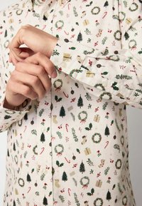 Mani che regolano il polsino di una camicia bianca con bottoni, decorata con un motivo natalizio ripetuto composto da alberi, ghirlande, regali, bastoncini di zucchero e calze.