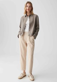 Lichtgrijs knoopjack over een wit shirt, gecombineerd met gestructureerde beige broek en beige sneakers met bruine accenten. Eenvoudig design en pasvorm.