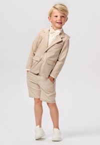Beige colbert met twee zakken en een enkele knoop, gecombineerd met bijpassende shorts. Lichtgekleurde shirt en vlinderdas. Witte sneakers.