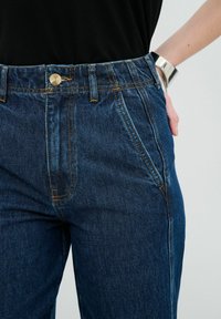 Tmavě modré džíny s vysokým pasem, se žlutým kontrastním prošíváním, bočními kapsami a stříbrným zapínáním na knoflík.