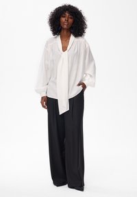 Biała bluzka z długim rękawem, z wiązaniem pod szyją i szerokimi mankietami, zestawiona z czarnymi spodniami z wysokim stanem i plisami, ukazująca gładkie faktury i dopasowany krój.