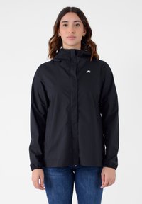 Giacca leggera nera con cappuccio, dotata di polsini elastici, zip frontale e un piccolo logo sul petto, indossata sopra a jeans blu.