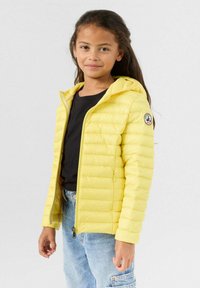 Geel gewatteerd jack met capuchon, voorzien van doorgestikte panelen en een logopatch op de mouw. Draagt over een zwart shirt en spijkerbroek.