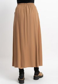Brauner Maxikleid mit elastischem Bund, glatter Textur, seitlichen Taschen und fließendem Design, kombiniert mit schwarzen Socken und Schuhen.