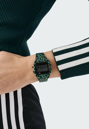 Digitale horloge met een groene rubberen band met luipaardprint, rechthoekige zwarte wijzerplaat met tijdsaanduiding, gedragen om een pols met een gestreepte mouw.