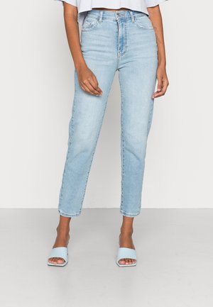 Kobieta w jasno-niebieskich dżinsach z wysokim stanem i jasno-niebieskich mule na obcasie z odkrytymi palcami stoi na tle jednolitego tła.