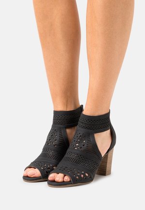 Sandales à talons noirs découpées avec motifs géométriques. Fabriquées en matériau synthétique texturé, avec un talon épais en bois et un design à bout ouvert.