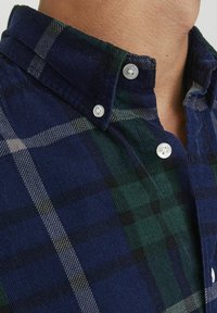 Camisa de botones con un patrón de cuadros en azul y verde, hecha de una tela texturizada con un cuello clásico y botones de plástico blanco.