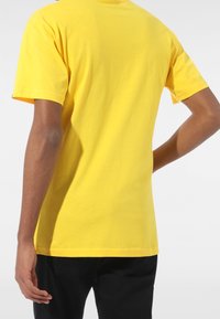 Camiseta de algodón amarilla con mangas cortas, escote redondo y un corte relajado, vista desde la parte de atrás con pantalones cortos negros visibles por debajo.