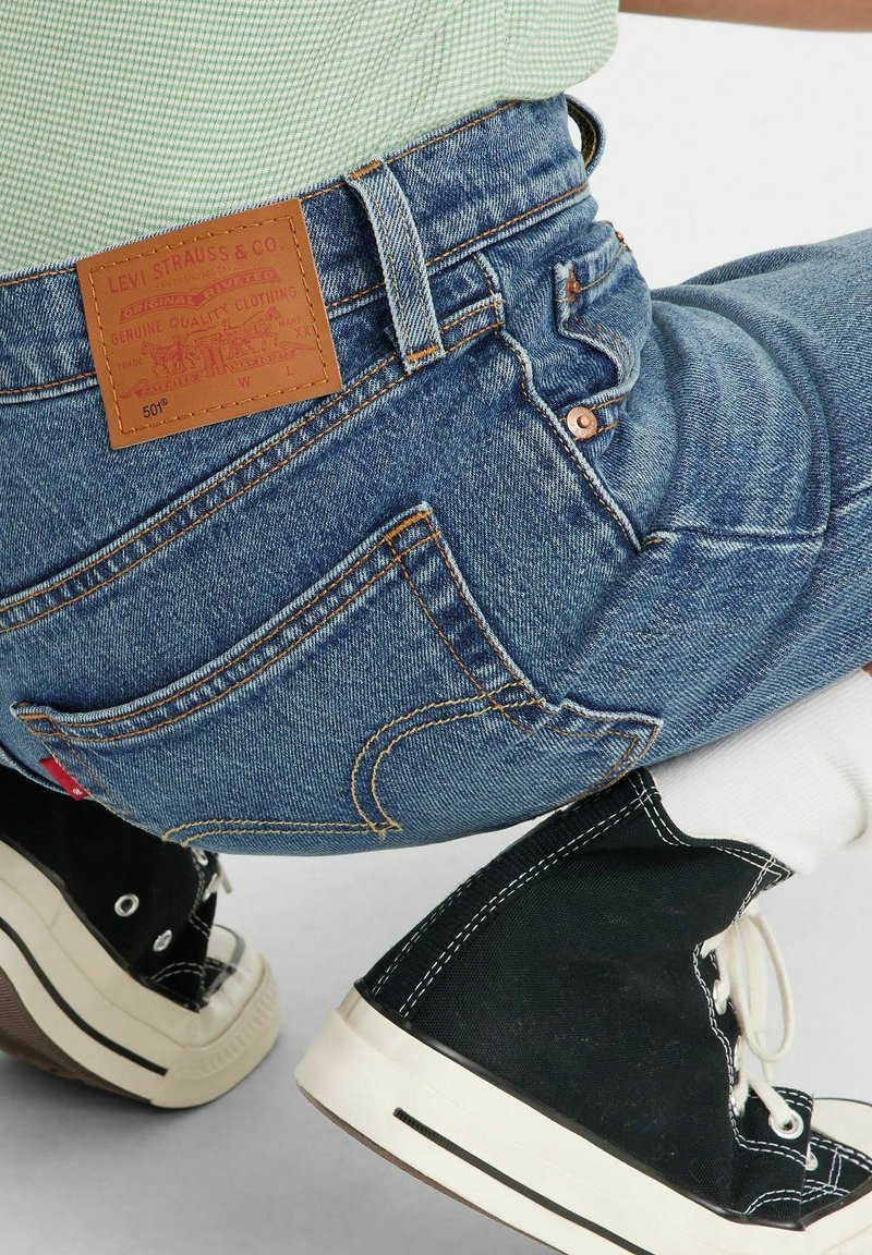 Jeans en denim bleu foncé avec une étiquette en cuir marquée "LEVI STRAUSS & CO." et des coutures beiges. Baskets montantes noires avec des lacets et des semelles blanches.
