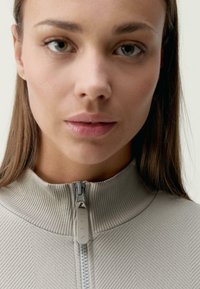 Texturovaný, světlě šedý zip-up oděv s vysokým límcem a centrálním zipem. Výrazným prvkem je tahací zip s jemným logem.