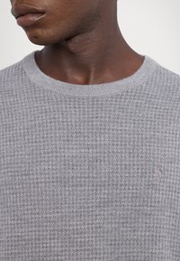 Gros plan d'une personne portant un pull en maille texturée gris clair avec un col rond et un petit logo brodé.