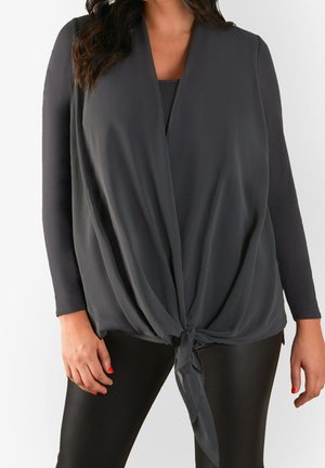 Dunkelgraues, leichtes Oberteil mit drapiertem Vorderteil, V-Ausschnitt und Bindedetail in der Taille. Lange Ärmel kombiniert mit schwarzen Leggings.