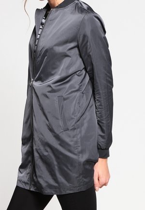 Frau trägt eine lange, glänzende dunkelgraue Bomberjacke mit Seitentaschen und Frontreißverschluss, kombiniert mit schwarzen Hosen.