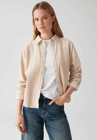 Woman in beige corduroy button-up shirt over white t-shirt and blue jeans, standing with one hand in pocket against plain background.
