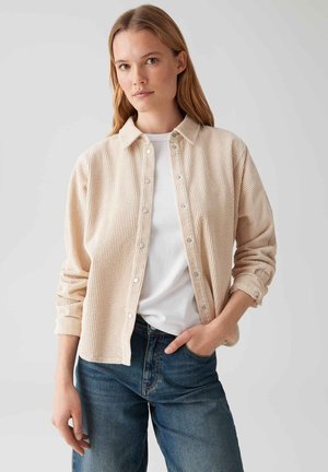 Femme portant une chemise en velours côtelé beige à boutons sur un t-shirt blanc et un jean bleu, se tenant avec une main dans la poche contre un fond uni.