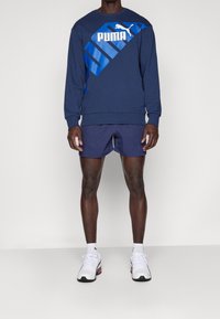 Sudadera azul marino con un diseño diagonal en azul y el logo blanco de "PUMA", combinada con pantalones cortos azul marino y zapatillas blancas. Fondo liso.