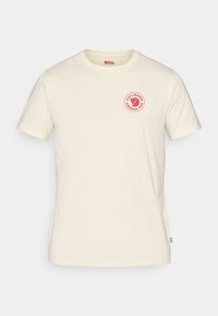 Krémové bavlněné tričko s krátkým rukávem, které má na hrudi kruhové červené logo a malou vlajkovou aplikaci na boku.