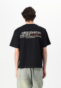 Schwarzes T-Shirt mit kurzen Ärmeln, das auf der Rückseite mit weißem und rotem Text versehen ist. Es hat eine lockere Passform und eine glatte Baumwolltextur. Kombiniert mit hellgrünen Jeans.