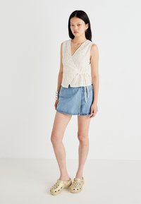 Wrap-stil kremetop med teksturt mønster, parret med en lyseblå denimskjørt. Modellen har på seg gullklokker med dekorative detaljer.
