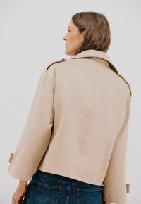 Beige trenchcoat med bred krage och trekvartslånga ärmar. Slät textur med en strukturerad silhuett och knappar på axlarna.