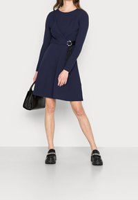 Mörkblå klänning med långa ärmar, twist-front design och silverringdetalj vid midjan. Matchad med en texturerad svart handväska och träskor.