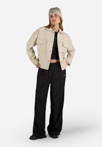 Giacca beige cropped con grandi tasche frontali, top nero corto e pantaloni neri morbidi. Berretto beige a maglia testurizzata con dettaglio del logo. Scarpe nere.
