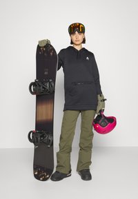 Snowboard s tmavým drevom a čiernym dizajnom, čierne viazanie; osoba oblečená v čierneho mikiny, olivovo zelených nohaviciach a ružovej helme.
