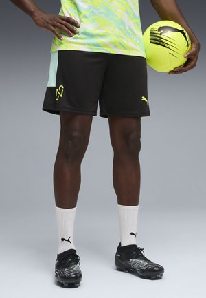 Persona che indossa pantaloncini da calcio neri Puma, calzini bianchi Puma, scarpe chiodate nere, che tiene una palla da calcio gialla e nera su uno sfondo semplice.