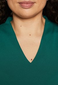 Bluzka w kolorze turkusowym z dekoltem w serek i fakturowanym materiałem. Posiada gładki kołnierzyk i widoczną skórę, w tym pieprzyki na szyi.
