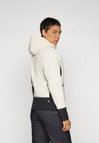 Lehká prošívaná bunda v krémové barvě s kapucí, se zapínáním na černý zip v oblasti pasu a texturovanými panely. Sportovní a přiléhavý design.
