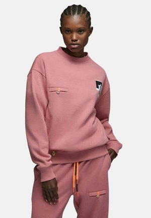 Femme portant un sweat-shirt rose ample assorti et un pantalon de survêtement avec petites poches boutonnées et cordons orange, posant devant un fond uni.