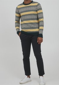 Musterstrickpullover mit blauen, gelben und weißen Designs, gerippte Bündchen und Saum, kombiniert mit schwarzen Hosen und weißen Sneakern.