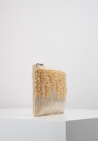 Kopertówka z koralikami w kremowym materiale, z teksturowanym frędzlem z złotych koralików, zamknięciem na suwak oraz o kształcie prostokątnym i sztywnej formie.