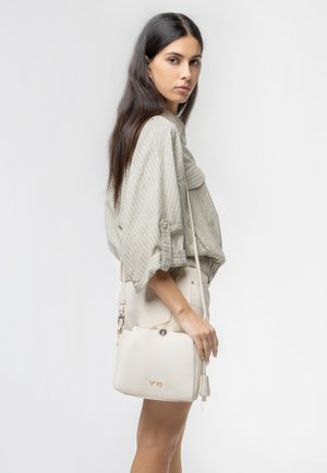 Giovane donna con lunghi capelli scuri, in camicetta leggera a righe e shorts beige, che tiene una borsa a tracolla color crema su sfondo bianco.