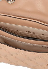 Beige gesteppte Leder-Handtasche mit einem Reißverschlussfach und einer Innenauskleidung, die mit dem Markennamen "GUESS" in Braun bedruckt ist.