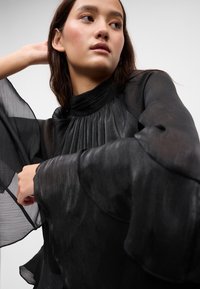 Mustmust muströöpidega pluus kõrge kaelusega ja laiade, voolavate varrukatega. Valmistatud kergest, tekstuurist kangast, millel on peen sära.