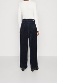 Námořnické kordové široké kalhoty s vysokým pasem, texturovanými vertikálními pruhy, dvěma zadními kapsami a jemnou logo nášivkou. V kombinaci s bílým topem s dlouhým rukávem.