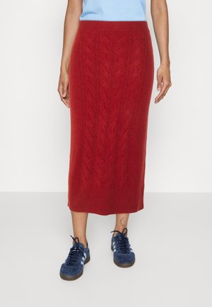 Person trägt einen roten, kabelgestrickten Midirock mit einem hellblauen Oberteil und marineblauen Sneakers mit weißen Streifen.