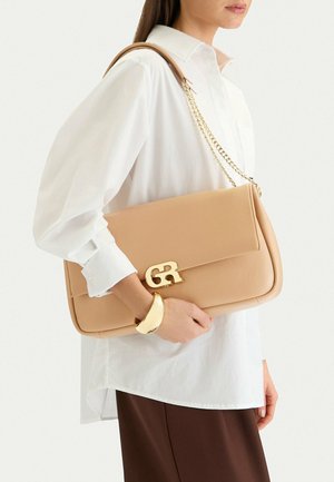 Donna con camicia bianca e gonna marrone tiene una borsa in pelle color cuoio con catenella dorata e chiusura con logo "GR" dorato.