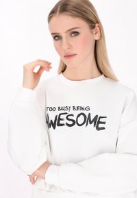 Fehér pulóver fekete szöveggel "TÚL FOGLALTAN KIVÁLÓNAK LENNI", kerek nyakkivágású, hosszú puffos ujjakkal, sima anyagból és bordázott mandzsettával.