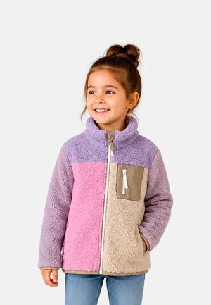 Jeune fille souriante, portant une veste polaire bicolore avec des sections violet, rose et beige, debout devant un fond clair uni.