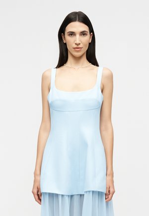 Femme aux cheveux bruns raides, portant une robe bleu clair sans manches et un délicat collier de perles, de face sur un fond uni.