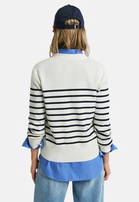 Pull rayé crème avec des rayures navy, porté sur une chemise bleu clair. Il présente des manches longues et un ourlet côtelé. Style décontracté et superposé.