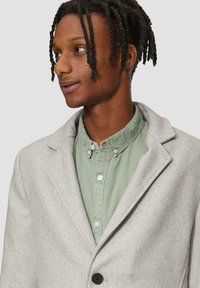 Lichtgrijze wolmix jas met notch-lapels en enkele knoopsluiting, gelaagd over een groene overhemden met knopen. Gelaagd afwerking en op maat gemaakte pasvorm.