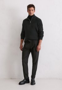 Pull en maille vert foncé avec col zippé demi, associé à un pantalon noir et des mocassins. Présente des poignets côtelés et une finition texturée.