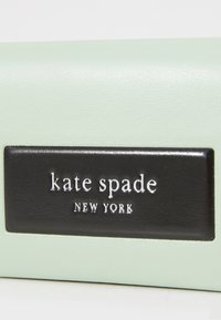 Matériau texturé vert menthe avec une étiquette rectangulaire noire embossée portant "kate spade NEW YORK" en lettrage blanc. Design simple et moderne.