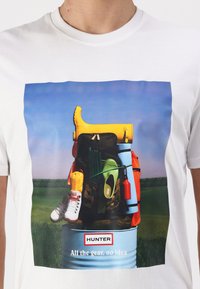 White t-shirt featuring a graphic print of various outdoor gear including yellow wellies, green boots, an orange backpack, and a blue barrel.