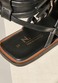 Must musta sandaalid lame taldaga, millel on rihmad ja pruun aktsent põhjas. Kuldne logo tekst ütleb "N. PREMIUM COLLECTION."