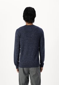 Suéter azul marino con un tejido texturado, escote redondo y puños y dobladillo de canalé. Usado con pantalones gris claro, visto desde atrás.